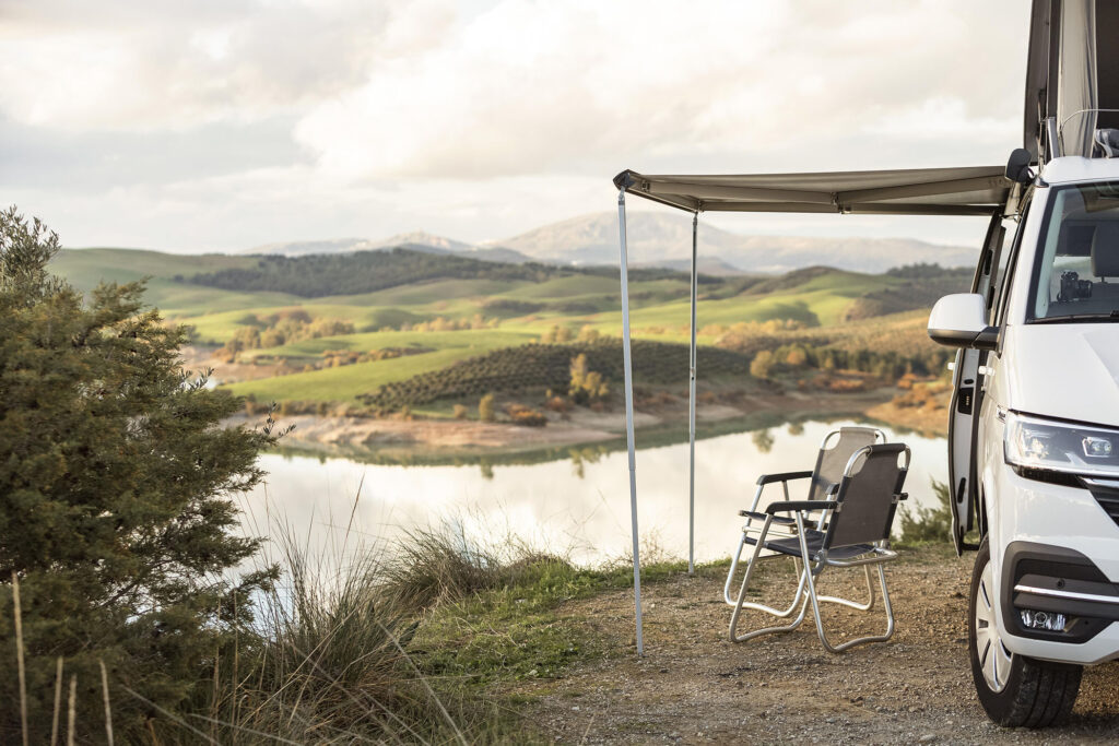Weisser Campervan mit ausgefahrener Markise und Campingstühlen an einem See – Hitzeschutz und Klimaanlage im Wohnmobil sind hier entscheidend