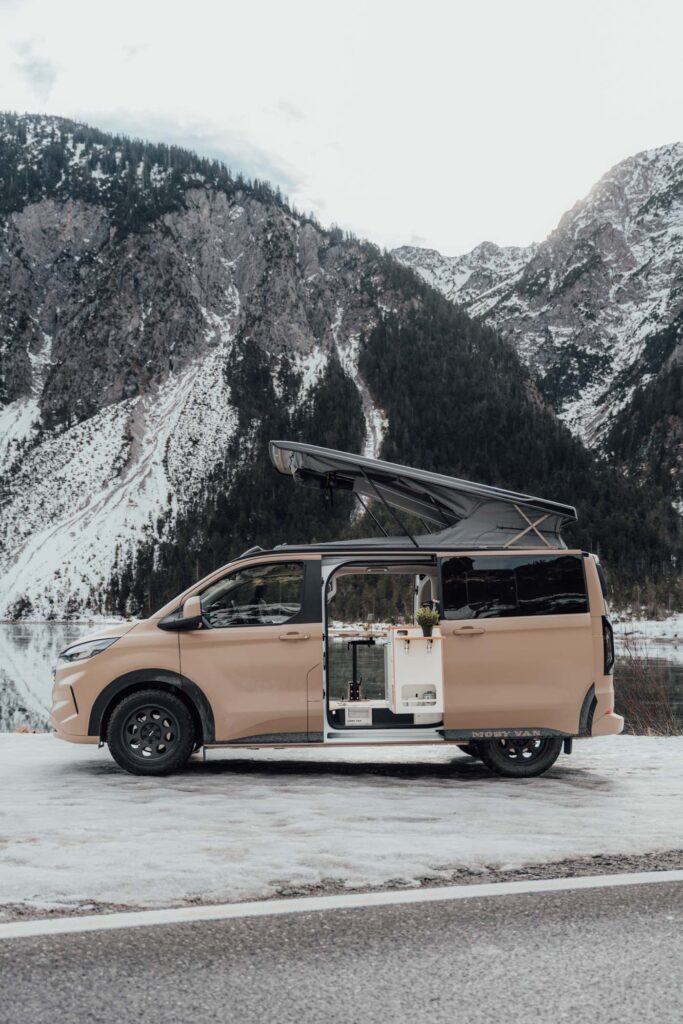 MOBY VAN Fordinand vor winterlicher Bergkulisse mit See – aufgestelltes Dach und offene Schiebetür zeigen den Innenausbau, BiwakLab ist offizieller Schweizer Händler des MOBY VAN Fordinand